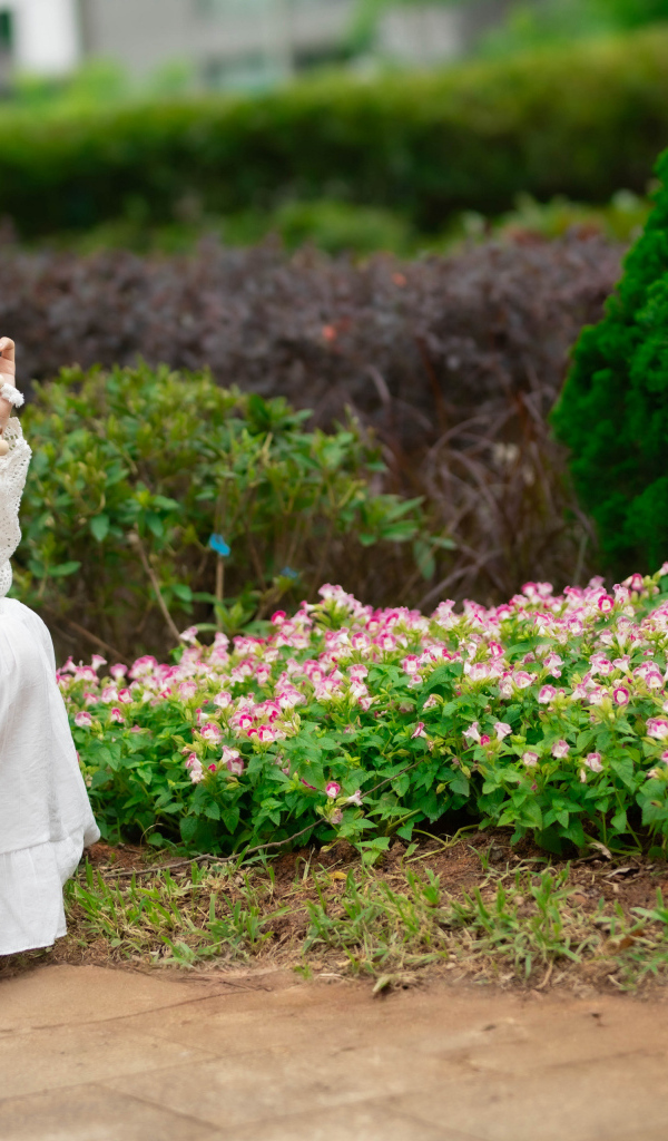 Милая азиатка в белом наряде сидит у клумбы с цветами