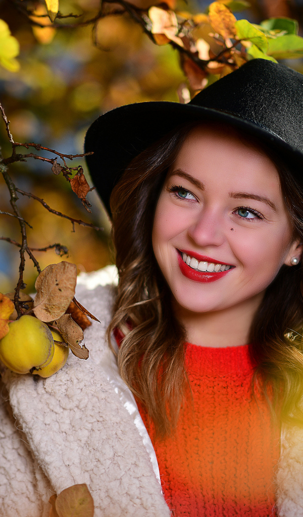 Улыбающаяся девушка в черной шляпе у дерева с яблоками
