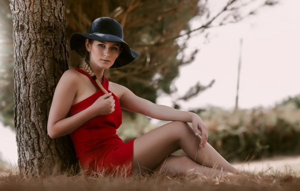 Красивая девушка в красном платье и черной шляпе сидит под деревом