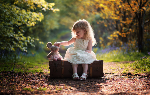 Маленькая девочка в белом платье сидит на большом чемодане с игрушкой