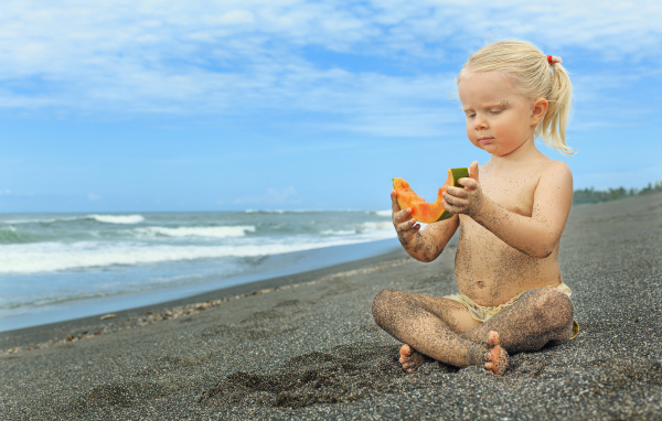 Маленькая девочка сидит на песке у моря с кусочком дыни