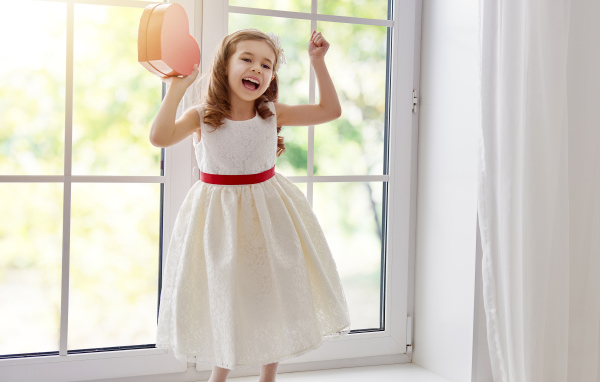 Маленькая девочка с коробкой в форме сердца в руках стоит на подоконнике