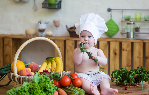 Маленький ребенок в шапке повара сидит на столе с овощами
