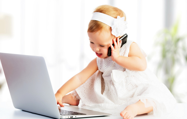 Маленькая девочка в белом платье сидит с телефоном у ноутбука