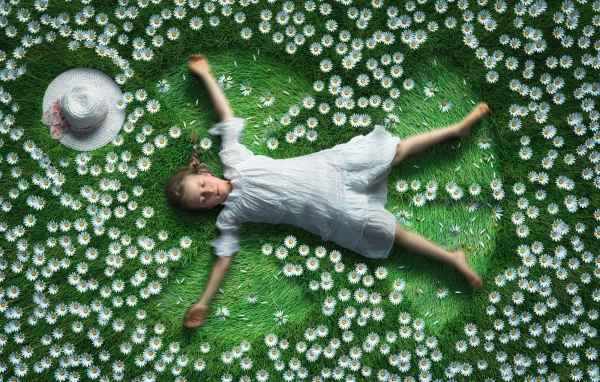 Маленькая девочка в белом платье лежит на зеленой траве с ромашками