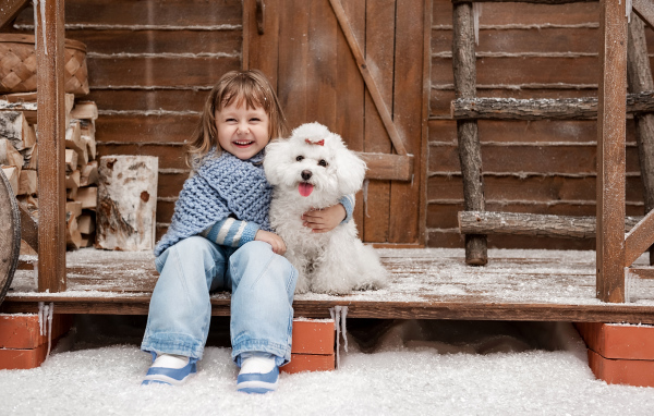 Маленькая улыбающаяся девочка сидит с белым пуделем на крыльце дома зимой
