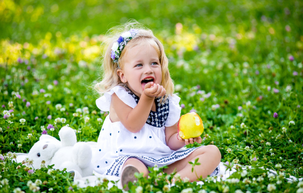Маленькая улыбающаяся девочка с яблоком сидит на зеленой траве