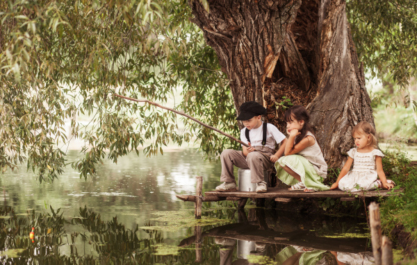 Две маленькие девочки и мальчик сидят на берегу пруда с удочкой