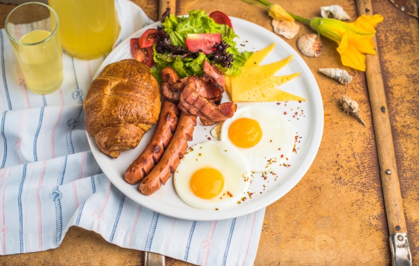 Яичница, салат, сосиски и круассан с соком на завтрак 