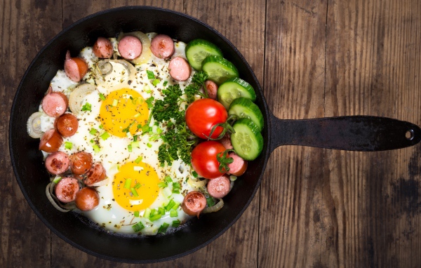 Яичница в сковороде с овощами и кусочками сосиски