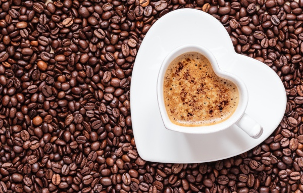 Белая чашка кофе в форме сердца стоит на кофейных зернах