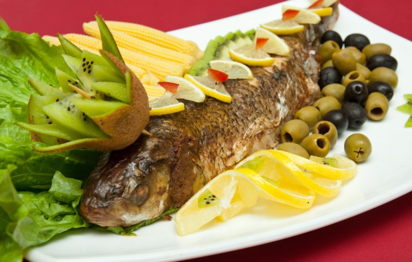 Запеченная рыба с оливками, лимоном, киви, кукурузой и листьями салата на белой тарелке