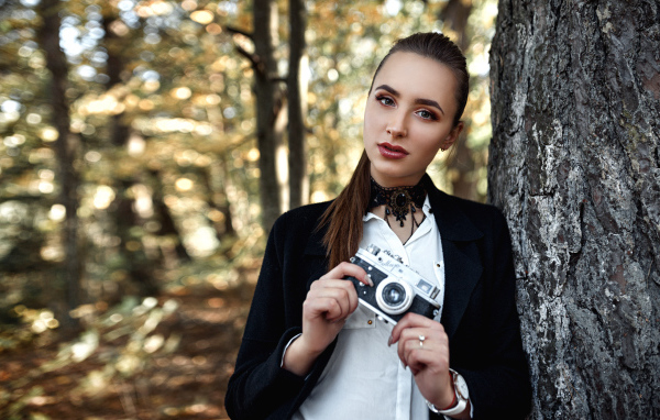 Красивая девушка с фотоаппаратом в руках стоит у дерева