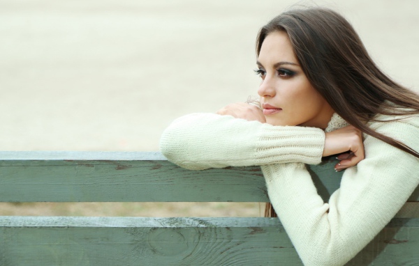 Задумчивая девушка брюнетка в белом свитере сидит на лавке