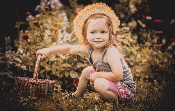 Маленькая девочка в шляпе сидит на траве с корзиной