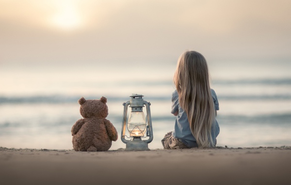 Маленькая девочка сидит на песке с плюшевым мишкой 