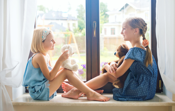 Две маленькие девочки с игрушками сидят на подоконнике у окна