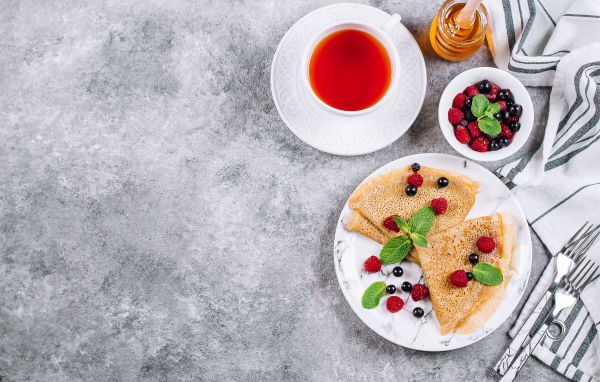 Тонкие блинчики на столе с ягодами, чаем и медом