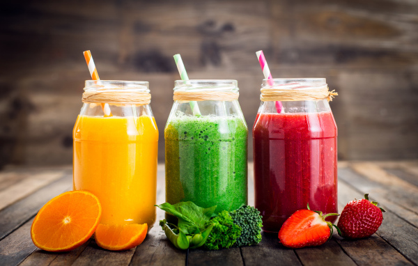 Три банки смузи с трубочкой на столе с апельсином, брокколи и клубникой