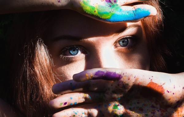 Красивая голубоглазая девушка с руками в разноцветной краске у лица