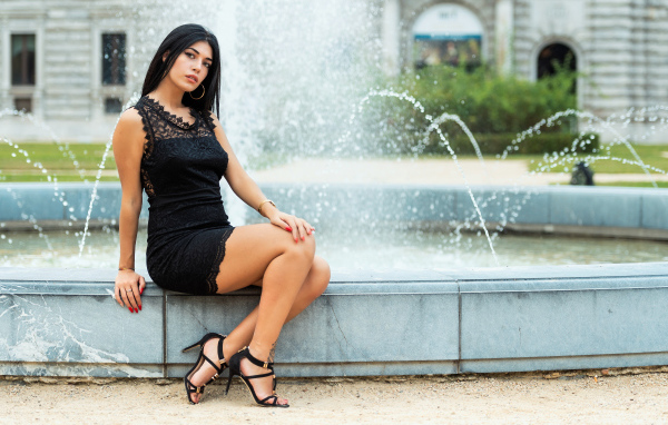 Красивая брюнетка в черном платье сидит у фонтана