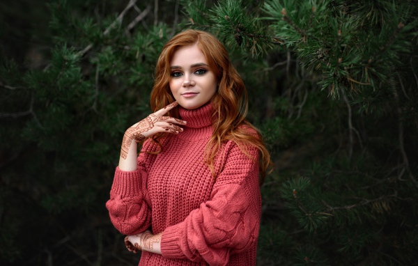 Рыжеволосая девушка в розовом свитере стоит у ели