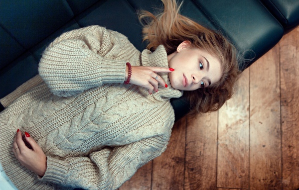 Молодая девушка в вязаном свитере лежит на черном кожаном диване