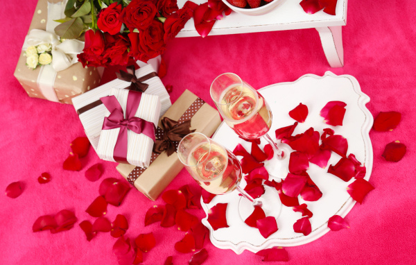 Два фужера с шампанским на красном покрывале с лепестками, подарками и букетом роз для любимой