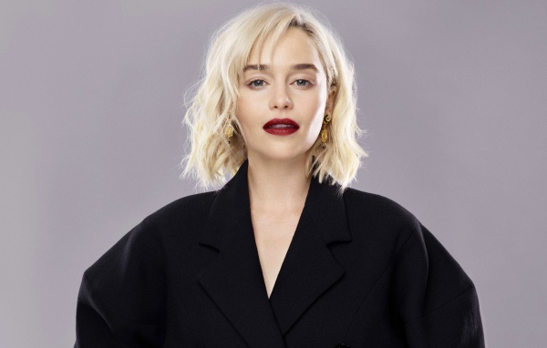 Яркая блондинка, актриса Эмилия Кларк в черном пальто на сером фоне