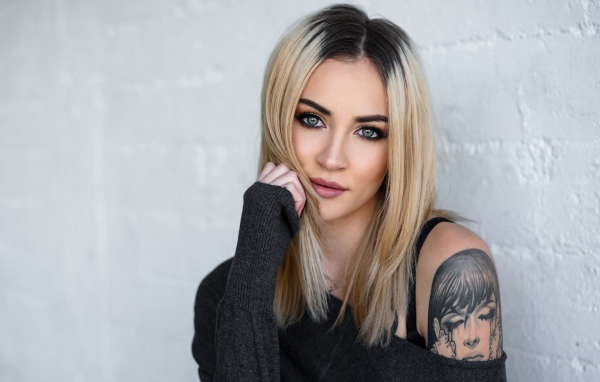 Стильная блондинка с татуировкой на плече на фоне стены