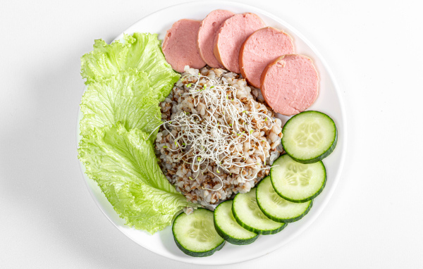 Гречка на тарелке с огурцом, колбасой и листьями салата на белом фоне