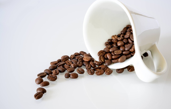 Кофейные зерна высыпают из белой чашки на стол 