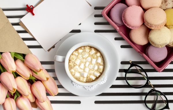 Чашка какао с маршмеллоу на столе с букетом тюльпанов, десертом макарун и очками