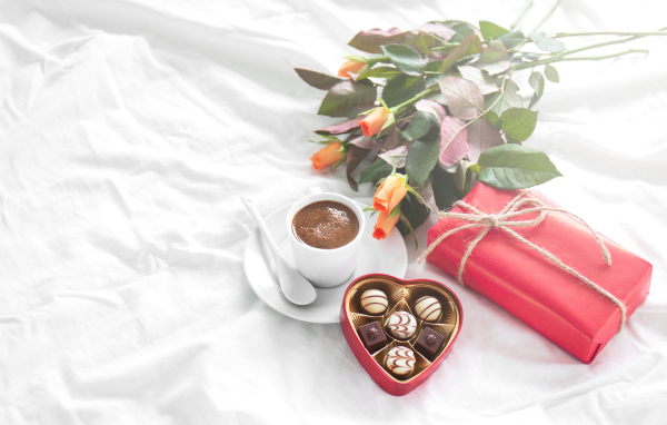 Чашка кофе на белой кровати с конфетами, подарком и букетом роз