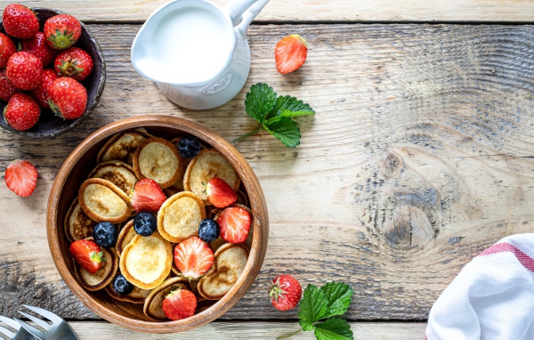 Маленькие оладьи в миске с клубникой и черникой на столе с молоком 