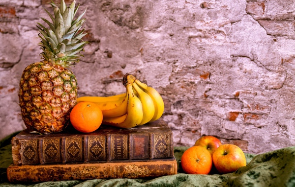 Ананас, бананы, апельсин и яблоки лежат на столе с книгами