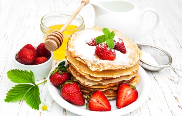 Тонкие блины с сахарной пудрой и ягодами клубники на столе с молоком и медом