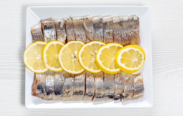 Селедка нарезанная кусочками на белой тарелке с лимоном 