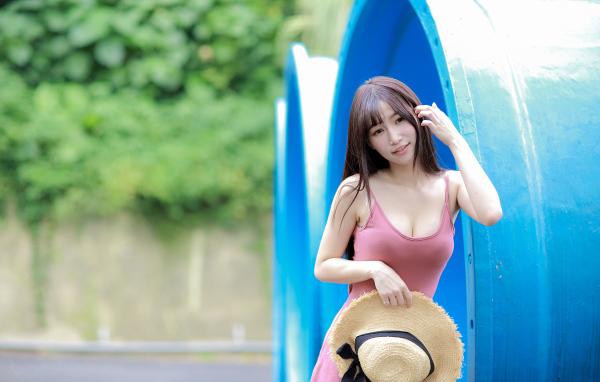 Стройная азиатка в платье со шляпой в руке стоит у синих  труб 