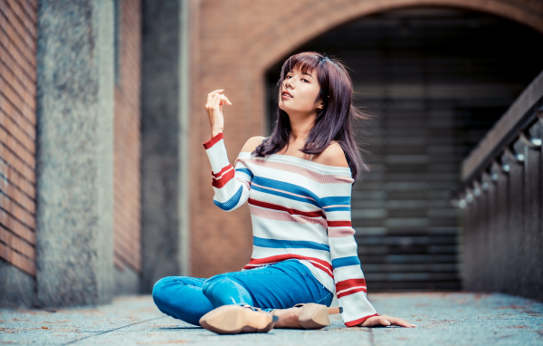 Девушка азиатка в джинсах и полосатом свитере сидит на асфальте