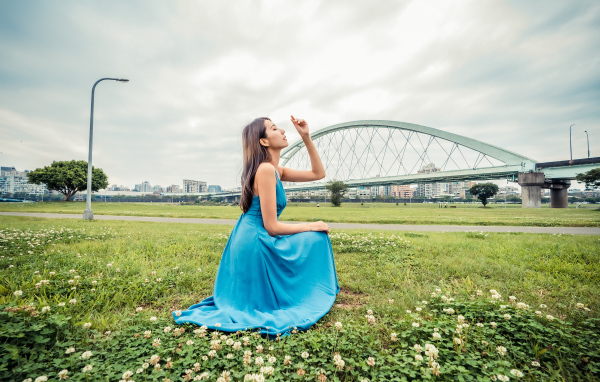 Красивая девушка азиатка в голубом платье  сидит на траве на фоне моста