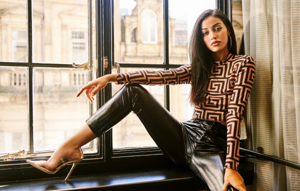 Красивая брюнетка в черных брюках сидит на окне 