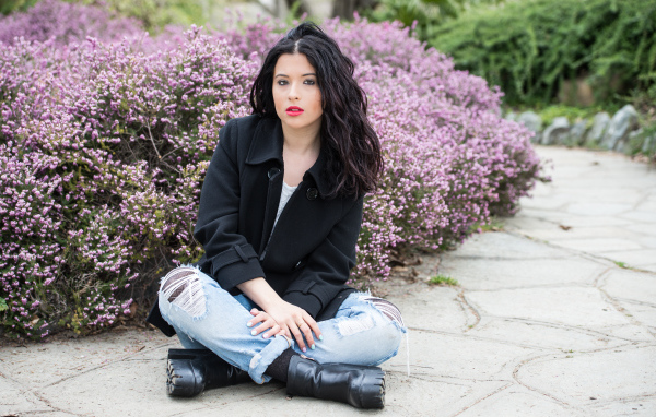 Красивая девушка в черном пальто сидит на земле у кустов