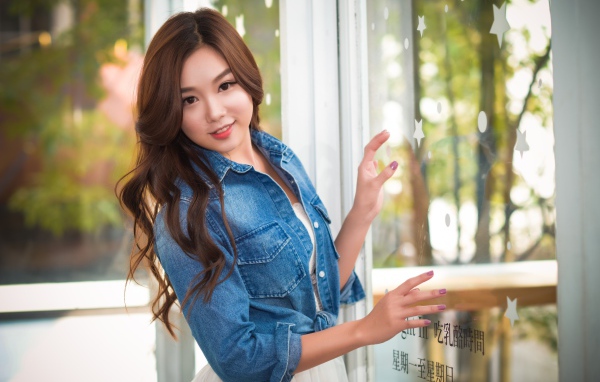 Милая девушка азиатка в джинсовой куртке стоит у окна 