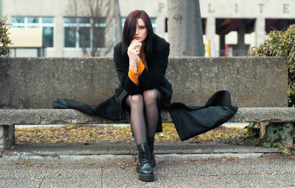 Девушка в черном пальто сидит на бетонной лавке 