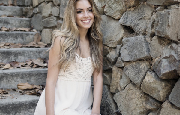 Улыбающаяся  девушка в белом платье стоит на ступеньках у стены