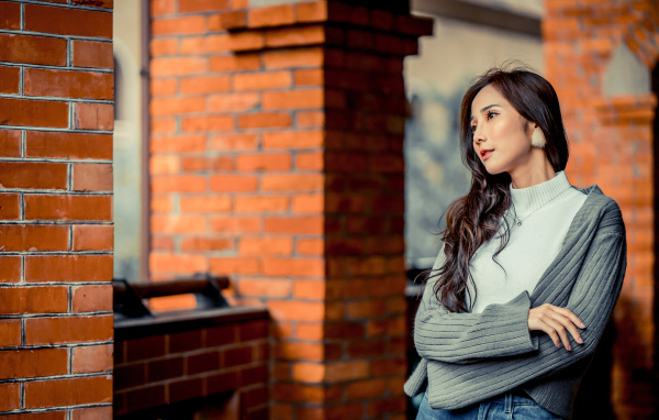 Молодая девушка азиатка в серой кофте у кирпичной стены 
