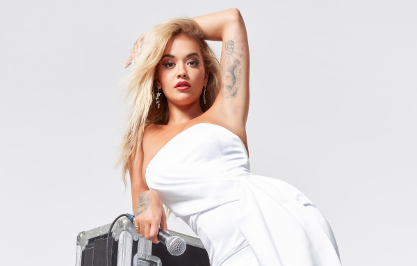 Певица Рита Ора в белом платье с микрофоном на сером фоне 