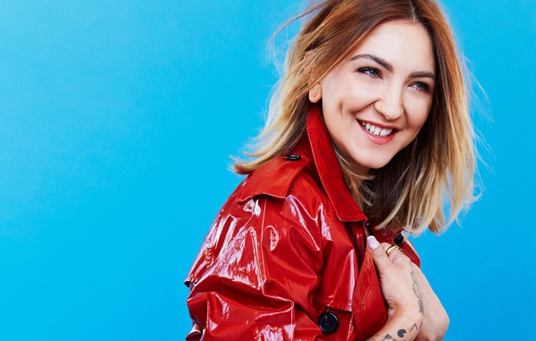 Улыбающаяся певица Джулия Майклз в красной куртке на голубом фоне