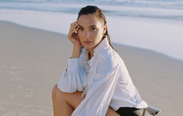 Актриса Галь Гадот в белой рубашке сидит на песке